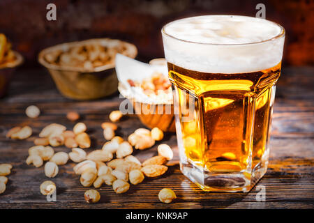 Glas mit frisch gezapftes helles Bier in der Nähe von Platten mit Snacks und zerstreut Muttern auf dunklem Holzschreibtisch. Nahrungsmittel und Getränke Konzept Stockfoto