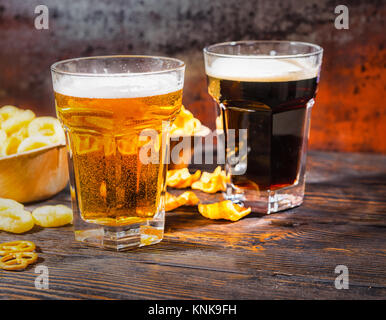 Zwei große Gläser mit frisch gezapftes helles und dunkles Bier in der Nähe von Platten mit Snacks und Chips auf dunklem Holzschreibtisch. Nahrungsmittel und Getränke Konzept Stockfoto