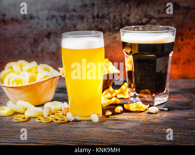 Zwei große Gläser mit frisch gezapften Licht ungefiltert und dunkles Bier in der Nähe von Platten mit Snacks und Chips auf dunklem Holzschreibtisch. Nahrungsmittel und Getränke Konzept Stockfoto