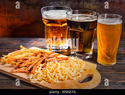 Drei Gläser mit Licht, ungefilterten und dunkles Bier stehen in einer Reihe in der Nähe von Holz Schneidebrett mit Snacks auf dunklen Schreibtisch. Nahrungsmittel und Getränke Konzept Stockfoto