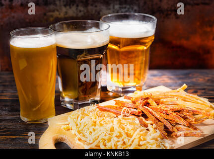 Drei Gläser mit Licht, ungefilterten und dunkles Bier stehen in einer Reihe in der Nähe von Holz Schneidebrett mit getrocknetem Fisch und Tintenfisch auf dunklen Schreibtisch. Nahrungsmittel und Getränke Stockfoto