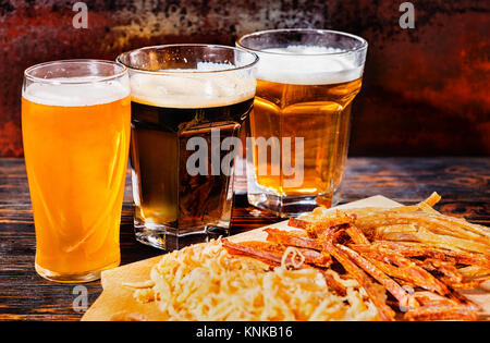 Drei Gläser mit Licht, ungefilterten und dunkles Bier stand in der Nähe von Holz Schneidebrett mit getrocknetem Fisch und Tintenfisch auf dunklen Schreibtisch. Nahrungsmittel und Getränke Konzept Stockfoto