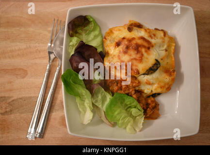 Essen Fotografie Bild von einem Teil der Köstlichen heißen hausgemachte Lasagne mit grünem Blattsalat in weißen Teller mit Besteck und einem hölzernen Hintergrund Stockfoto