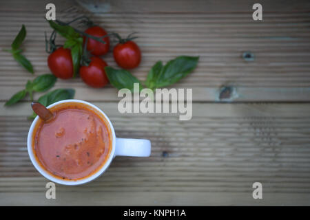 Essen Fotografie Bild der hausgemachten Tomatensuppe in Weiß Tasse oder Becher mit frischen Zutaten von Tomaten und Basilikum Blätter im Hintergrund auf Holz mit Platz Stockfoto