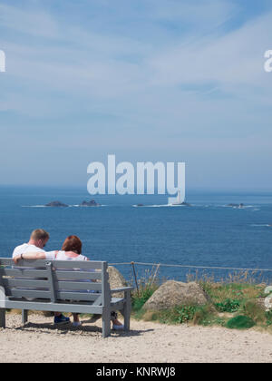 Junge kaukasier Paar auf einer Bank sitzen bei Land's End suchen, longships Leuchtturm, Penwith Halbinsel, Cornwall, England, Großbritannien im Sommer Stockfoto