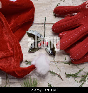 Nahaufnahme eines Santa Hut, ein paar gemütlichen roten Handschuhe, einige Zweige der Kiefern- und ein Zweig der Stechpalme auf einem weißen Holzmöbeln im Landhausstil Oberfläche Stockfoto