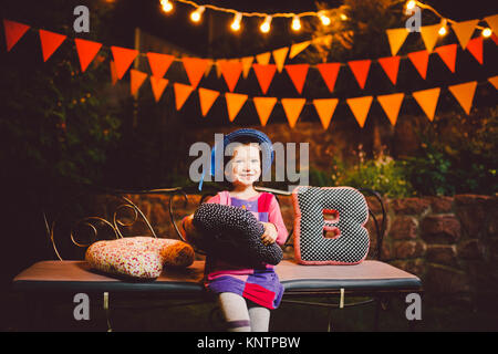 Ein lustiges Kind, das Mädchen sitzt auf einer Bank in einem festlich dekorierten Hof Einrichtung mit hellen Girlanden von Laternen. Sie lernt das Alphabet in spielerischer Form. Stockfoto