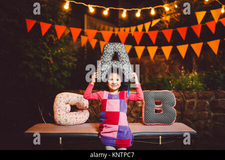 Ein lustiges Kind, das Mädchen sitzt auf einer Bank in einem festlich dekorierten Hof Einrichtung mit hellen Girlanden von Laternen. Sie lernt das Alphabet in spielerischer Form. Stockfoto