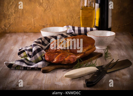 Horizontale Foto mit großen Stück geräuchertes Schweinefleisch Fleisch mit Knochen auf Schneidebrett platziert. Blau Handtuch, Häcksler, Gabel, Schüsseln und Rosmarin sind um mit w Stockfoto