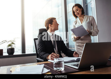 Zwei Geschäftsleute arbeiten zusammen im Büro. Geschäftsmann sitzt am Schreibtisch und seine Sekretärin liest Notizen in einem modernen Büro. Stockfoto