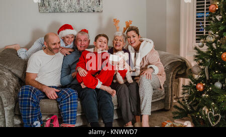 Drei generation Familie sind auf dem Sofa sitzen gemeinsam auf Weihnachten Morgen mit dem Haustier Hund. Sie sind alle in ihren Pyjamas und Nehmen ein selfie Stockfoto