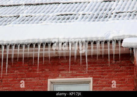 Weißer Schnee auf dem Dach eines roten Backsteinhaus und Eiszapfen hängen von einem Schieferdach. Stockfoto