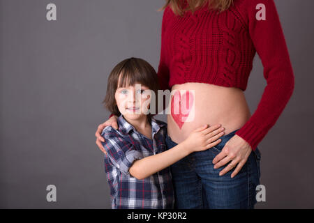 Schöne Vorschulkind, umarmt seine schwangere Mutter glücklich sein großer Bruder erwartet. Rotes Herz auf Mütter Bauch, Kind küssen ihr kleines b gezeichnet Stockfoto