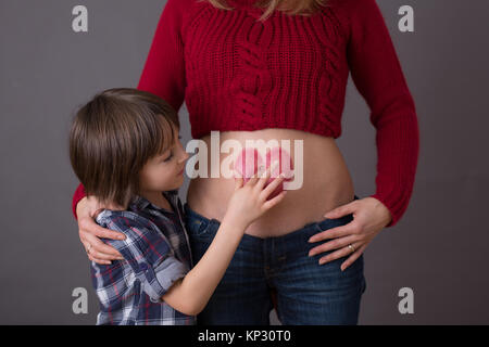 Schöne Vorschulkind, umarmt seine schwangere Mutter glücklich sein großer Bruder erwartet. Rotes Herz auf Mütter Bauch, Kind küssen ihr kleines b gezeichnet Stockfoto