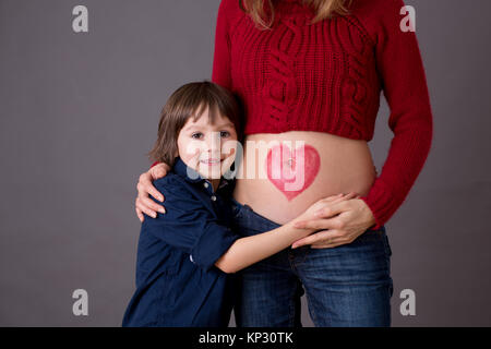 Schöne Vorschulkind, umarmt seine schwangere Mutter glücklich sein großer Bruder erwartet. Rotes Herz auf Mütter Bauch, Kind küssen ihr kleines b gezeichnet Stockfoto