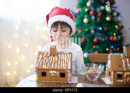 Cute little boy, die Lebkuchen cookies Haus, Dekorieren zu Hause vor dem Weihnachtsbaum, Kind spielen und genießen, Weihnachten Konzept Stockfoto