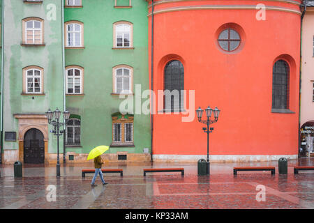 Man regen Stadt, ein Mann Unterschlupf unter einem gelben Regenschirm Wanderungen vorbei an einer Reihe von bunten Barockbauten in kleinen Marktplatz in Krakau, Polen. Stockfoto