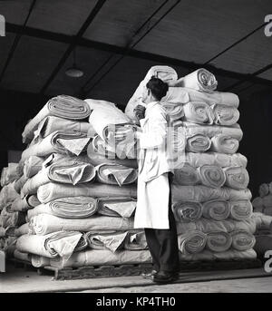 1950er Jahre, historische Bild, ein männlicher Arbeiter in weißen Overalls Kontrolle eine Rolle von Leinen unter den vielen, die ordentlich zusammen auf Paletten gestapelt werden, Nordirland. Stockfoto