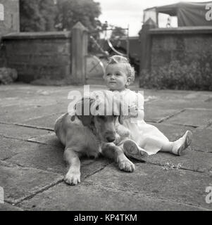 1950er Jahre, 'guten Gesellschaft'....historisches Bild, ein kleines Mädchen in einem Kleid und Schuhe gerne draußen auf einer Terrasse sitzen neben der Familie Haustier Hund, ein Labrador, der unten liegt, England, UK. Stockfoto