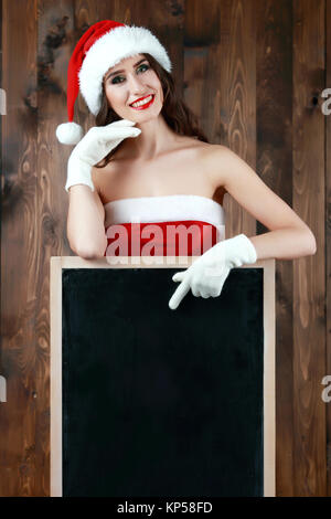 Hübsche junge Frau in santa claus Kleid und weiße Handschuhe holding Tafel über Holz- Hintergrund Stockfoto