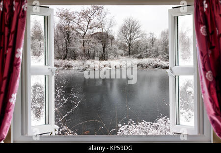 Blick durch die offenen Fenster auf einen schönen gefrorenen See winter schnee im ländlichen England. Rote Vorhänge hängen vor dem modernen Doppelt verglaste Fenster Stockfoto