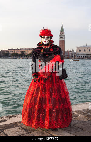 Karneval in Venedig, Venetien, Italien. Frau in markanten roten und schwarzen Kostüm an der Lagune mit dem Campanile und Dogenpalast hinter in Abend lig Posing Stockfoto