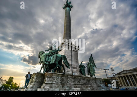 Ein junger Mann geht auf das Millenium Monument in Helden Platz, oder Hosok tere im City Park in Budapest Ungarn an einem bewölkten Nachmittag Stockfoto