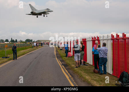 RAF Typhoon Jet Kampfflugzeug nähert sich der Landung bei Royal Air Force Coningsby mit Flugzeugbegeisterten und beobachtenden Beobachtern entlang des Zauns, über der Straße Stockfoto