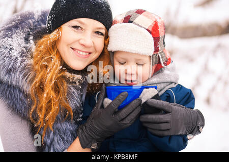 Baby mit junge schöne Mutter draußen im verschneiten Bäumen winter Hintergrund und trinken heißen Tee. Glückliche Familie genießen Sie einen wunderschönen Winter Tage auf Stockfoto