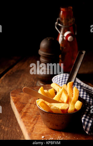 Knusprige Goldene Kartoffel Chips oder Pommes frites in einer rustikalen Metall Container auf einem Holz- Zähler mit einer Pfeffermühle und Ketchup für ein leckeres Mittagessen im Pub, mit Kopie Raum Stockfoto