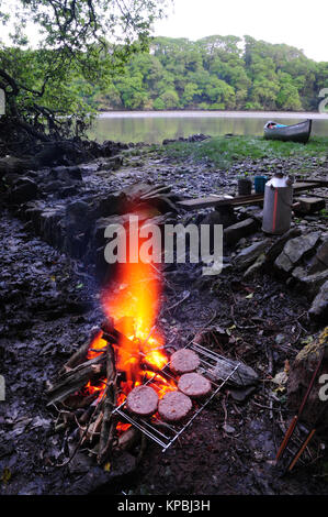 Kochen Rindfleisch Burger auf offenem Feuer während Kanu Camping. Im Hintergrund ist ein Kelly, für effiziente Wasser kochen, und ein Kanadier Kanu. Stockfoto