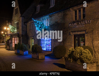 Bourton auf dem Wasser Geschäfte mit Weihnachtsbäumen und Lichter entlang der High Street in der Nacht. Bourton auf dem Wasser, Cotswolds, Gloucestershire, England Stockfoto