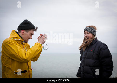 Mädchen gekleidet für den Winter mit bandana und Puffa-jacke, lacht wie ein Mann nimmt ihr Foto mit einer winterlichen Meer als Hintergrund, Cornwall. Stockfoto