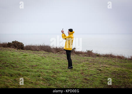 Mann in leuchtend gelben Jacke und schwarzen Hut steht am Rand einer Klippe, ein Foto mit seinem iPad, Rame Head, Cornwall. Stockfoto