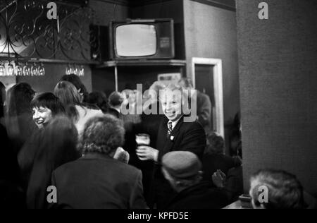 Londoner Pub aus den 1970er Jahren, voll mit Sonnentrinkern, Paar, die einen Witz teilen, lachen lächelnd. Ein Mann, der mit einem halben betrunkenen Pint Bier durch die Bar läuft. 70er London 1972 UK HOMER SYKES. Stockfoto