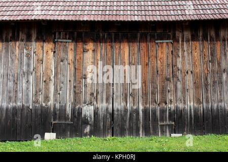 Große unlackierte Holz- scheune Türen mit starken Metallscharniere, roten Dachziegeln und ungeschnittenes Gras vor Auf warmen sonnigen Tag Stockfoto