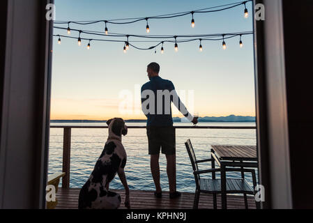 Fenster mit Blick auf den Menschen und Dogge Hund stehend auf seitlichem Meerblick Deck unter Hängeleuchten bei Sonnenuntergang gerahmtes Stockfoto