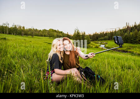 Zwei Freundinnen sitzen auf einem Grasplatz für ein selbst posiert - Porträt mit einem selfie Stick und Smart Phone bei Sonnenuntergang; Edmonton, Alberta, Kanada Stockfoto