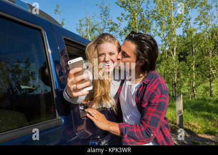 Ein junges Paar, ein Selbstporträt mit Ihrem Smart Phone auf Ihr Fahrzeug, während er sie auf die Wange küsst; Edmonton, Alberta, Kanada Stockfoto