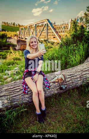 Eine junge Frau sitzt auf einem Baumstamm in einem Park ein Selbstporträt mit Ihrem Mobiltelefon mit einer Brücke und Fluss im Hintergrund; Edmonton, Alberta, Kanada Stockfoto