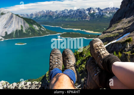 Close-up Wandern ein Paar Stiefel über eine Klippe mit Blick auf eine bunte alpinen See und Gebirge in der Ferne Stockfoto