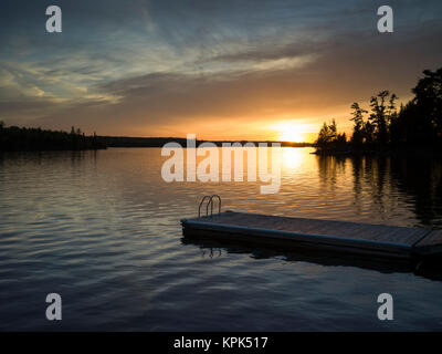 Die Sonne über eine ruhige See und Silhouette Bäume entlang der Küstenlinie mit einem Dock und Leiter in den Vordergrund Stockfoto