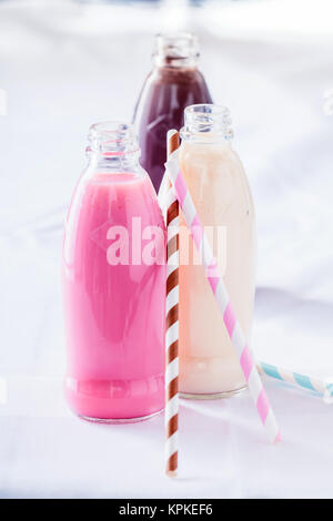 Drei große Flaschen voller Erdbeere, Schokolade und Vanille milchshakes Neben bunte Strohhalme auf weißen Tisch Stockfoto