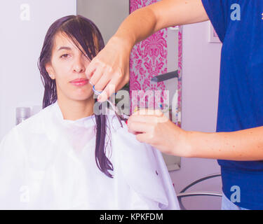 Frau mit langen dunklen nassen Haare mit Pony Haarschnitt im Friseursalon durch ein Stilist mit einer Schere. Die Frau ist mit einem weißen Cape abgedeckt. Stockfoto