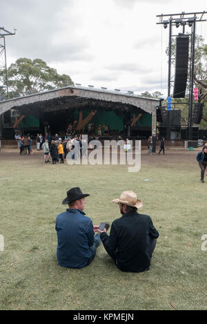 Zwei junge australische Kerle mit schwarzen und Stroh Cowboyhut sitzen auf Rasen vor der Musik Bühne sprechen. Stockfoto