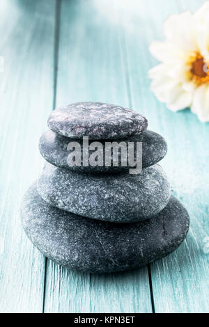 Gestapelte Glatte runde Steine für einen heißen Rock Spa Massage auf einem blauen Holzbrett Hintergrund mit Kopie Raum und eine frische weiße Blume in einem Wellness Konzept Stockfoto
