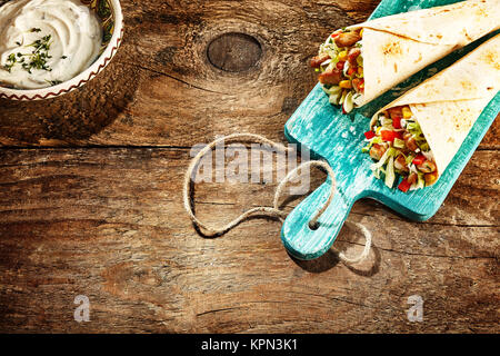 Hohen Winkel noch Leben auf Paar Chicken Fajitas gefüllt mit frischem Gemüse auf abgenutzten Blau Holz Schneidebrett auf rustikalen Holztisch mit Dip Sauce und reichlich Platz kopieren Stockfoto