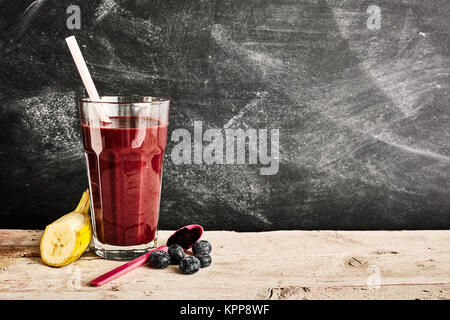 Hohes Glas des köstlichen Acai Berry Smoothie in zwischen einer Scheibe Banane, Blaubeeren und Plastik Löffel über Hintergrund mit schwarzen Brettes Platz kopieren Stockfoto