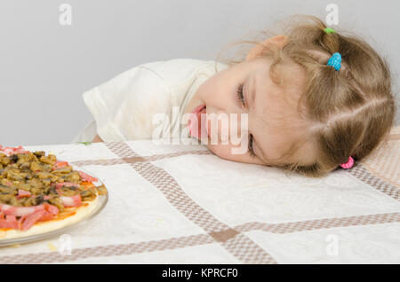 Kleines Mädchen mit hervorstehenden Zunge legte ihren Kopf auf den Tisch und schaut auf die pizza Stockfoto