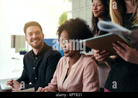 Positive junge afrikanische Geschäftsfrau am Schreibtisch in einem modernen Büro von lächelnden Kollegen umgeben Stockfoto